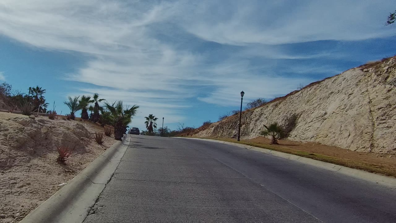 Forward road view in East Cape Los Cabos with gray asphalt, curb lines, palms, dry shrubs, rocky banks on both sides, distant streetlights, and no visible people.