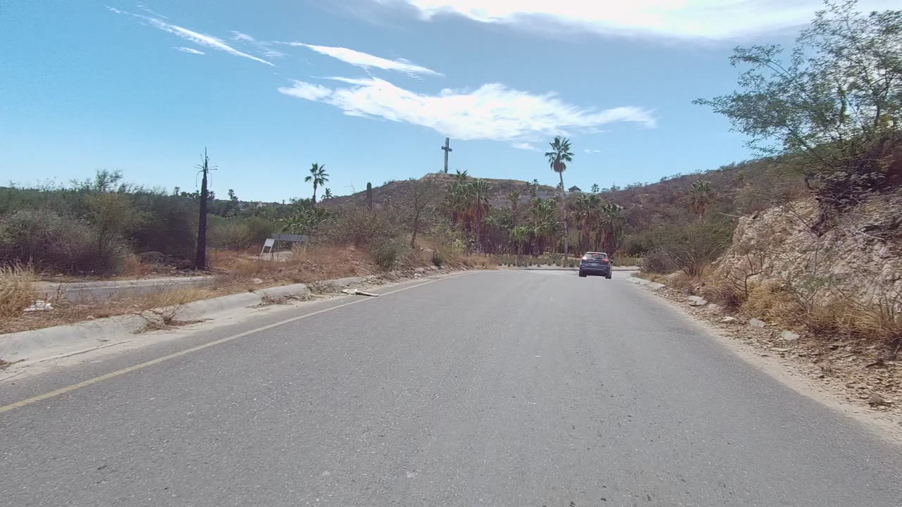 A dark car drives ahead on a paved road in East Cape Los Cabos, Mexico, with sandy shoulders, scrub, palms, a distant cross, dry hills, blue sky, and no visible people.