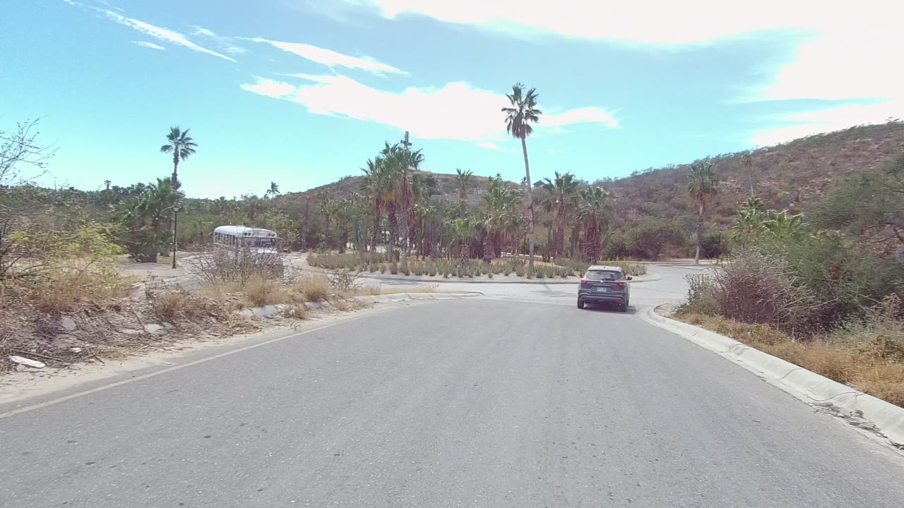 A white bus appears at the left side of the East Cape Los Cabos road junction as the dark car approaches, with palms, cacti, dry hillside, bright sky, and no visible people.