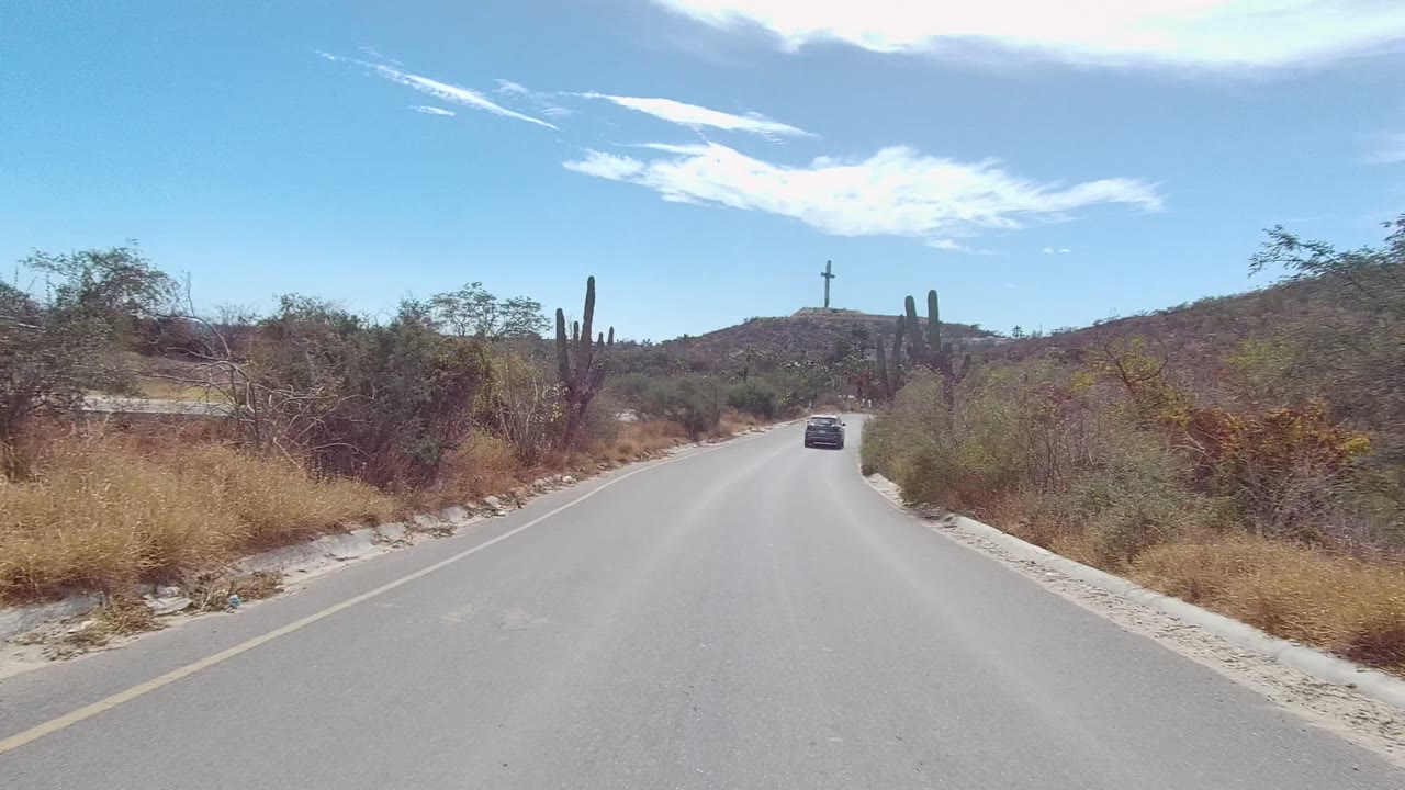 At 0:00, a paved road in East Cape Los Cabos, Mexico runs toward a small car ahead, with tall cacti, dry roadside grasses, low hills, scattered clouds, and no visible people.