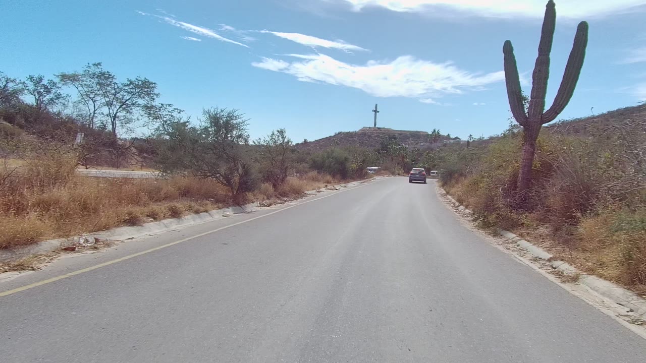 At 0:02, the road opens toward cactus-dotted hills in East Cape Los Cabos, with a large cactus on the right edge, dry brush on both shoulders, one car ahead, and no visible people.