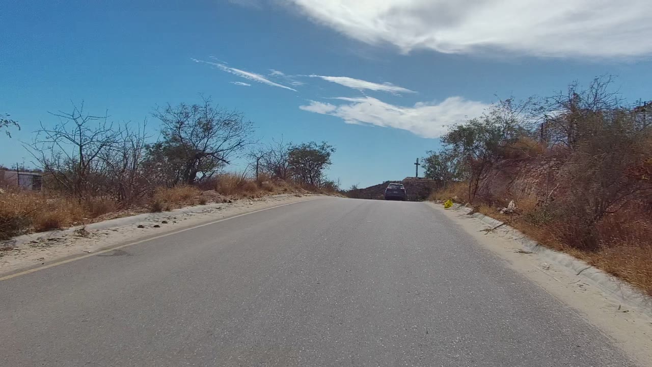 A paved road in East Cape Los Cabos, Baja California Sur runs through dry roadside brush toward a distant car under blue sky and white clouds, with no visible people.