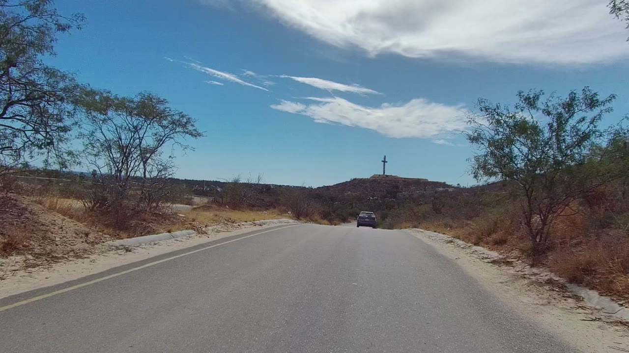 The paved road continues toward low hills in East Cape Los Cabos, with dry brush on both sides, a car ahead, and no visible people.