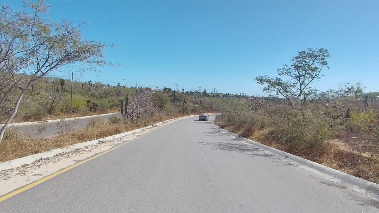 A paved road in East Cape Los Cabos, Baja California Sur curves through dry desert brush with cacti, low hills, and a distant car, with no visible people.