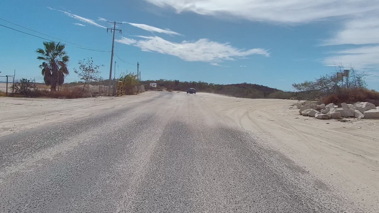 Opening view of an East Cape Los Cabos rural road with pale gravel, a palm at left, utility poles, scrubland, distant hills, blue sky, and no visible people.