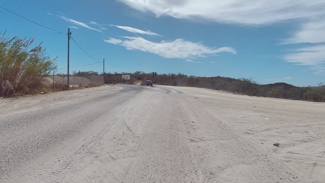 The East Cape Los Cabos road curves right past dry brush and a roadside sign, with a small car ahead, distant hills, bright blue sky, and no visible people.
