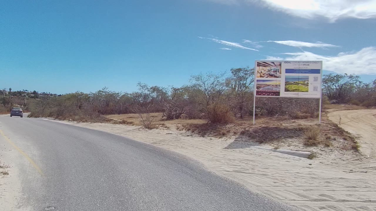 A small car drives away on the paved road in East Cape Los Cabos, passing a large roadside billboard, sandy shoulders, dry brush, blue sky, and no visible people.