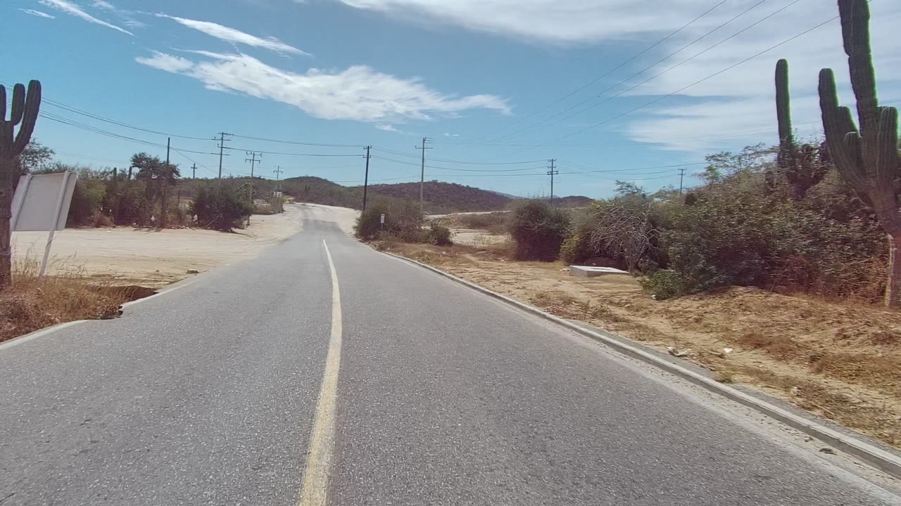 East Cape Los Cabos road view with the asphalt running straighter ahead between sandy shoulders, cactus stands, utility poles, sparse brush, hills, and no visible people.