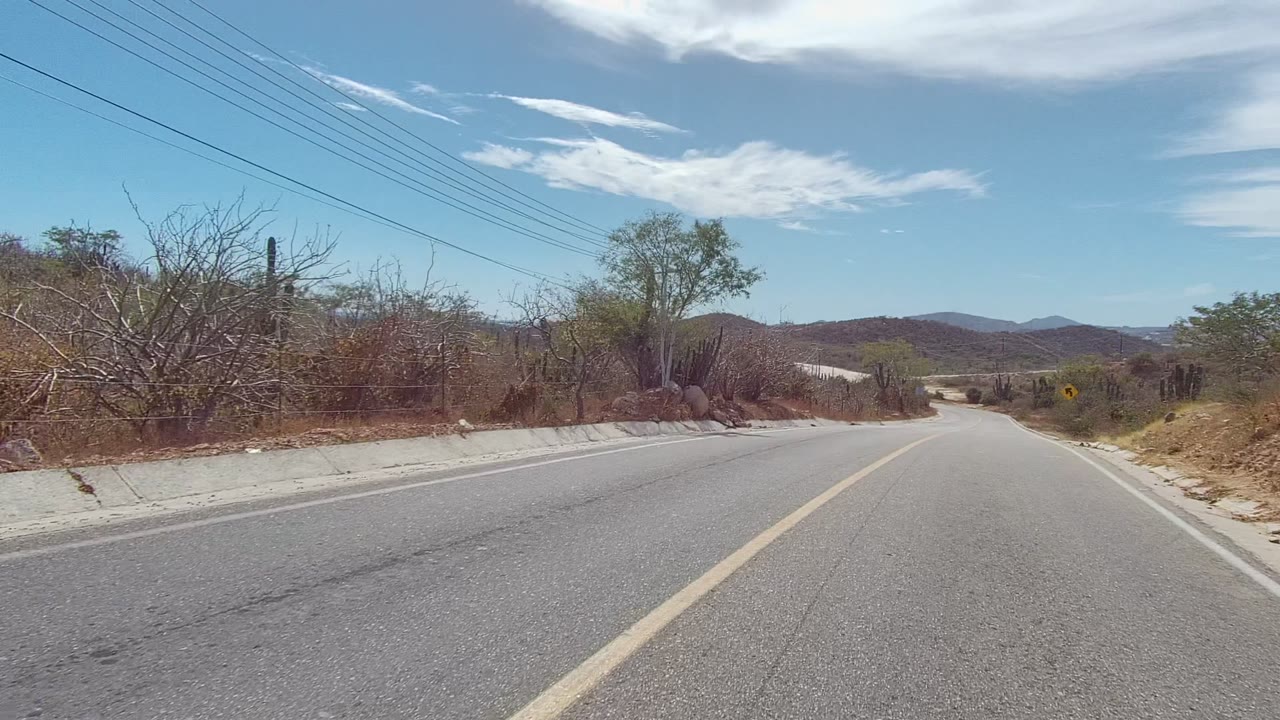 A two-lane road in East Cape Los Cabos, Baja California Sur, Mexico passes dry brush and cacti under a blue sky with no visible people.