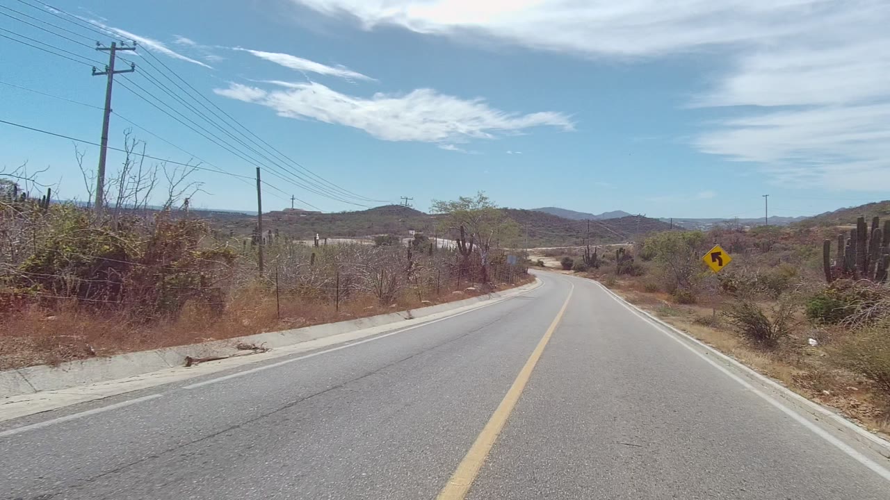 A paved road in East Cape Los Cabos bends through dry brush with a utility pole, roadside fence posts, a yellow curve sign, hills, and no visible people.
