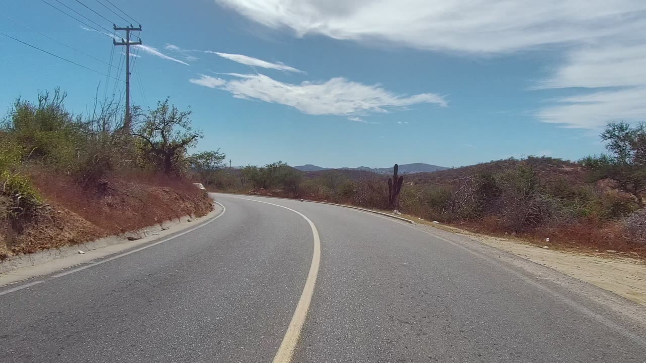 A curving paved road in East Cape Los Cabos, Mexico, passes dry brush, cactus, power poles, distant hills, and a blue sky, with no visible people.