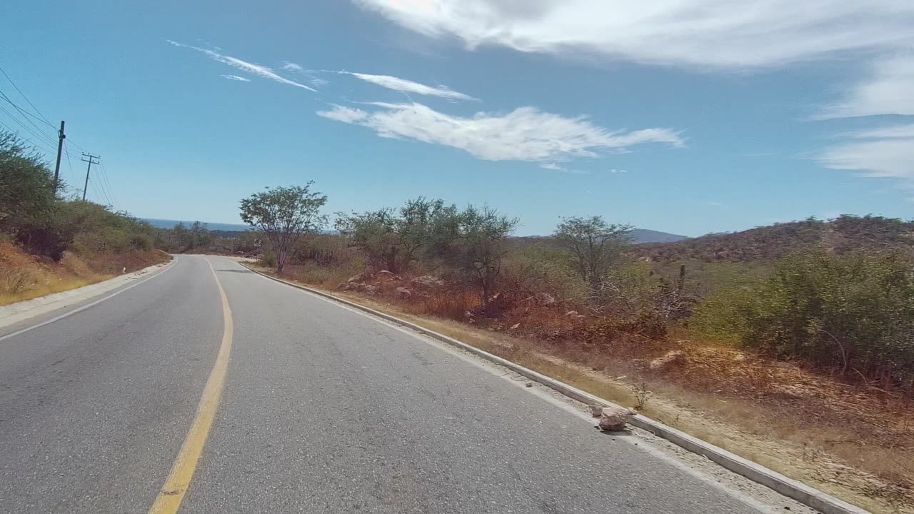 The road opens ahead in East Cape Los Cabos, Mexico, with cactus, scrub, dry roadside soil, low hills, and high white clouds under a clear blue sky, with no visible people.