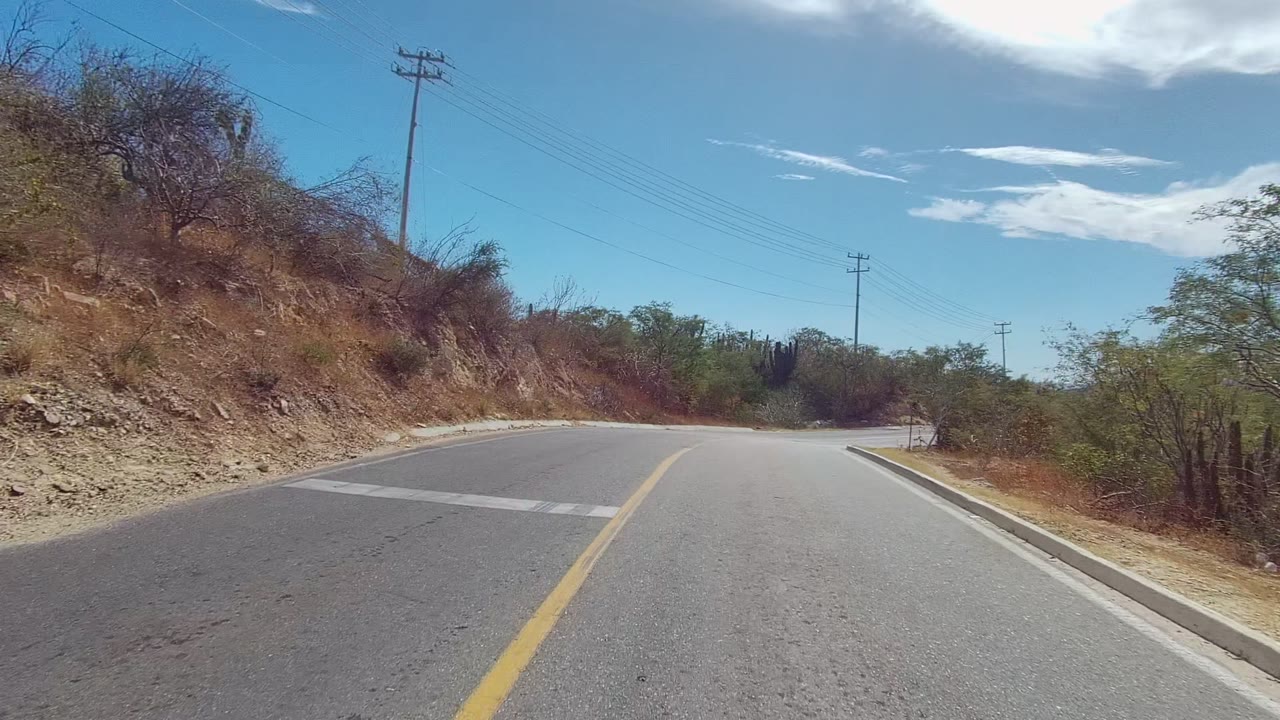 At 0:00, a paved road in East Cape Los Cabos, Baja California Sur curves ahead past dry brush, cactus, utility poles, and overhead power lines, with no visible people.