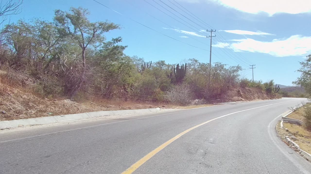At 0:02, the road in East Cape Los Cabos curves along dry brush and tall cacti, with a roadside guardrail edge, utility pole, power lines, distant hills, and no visible people.