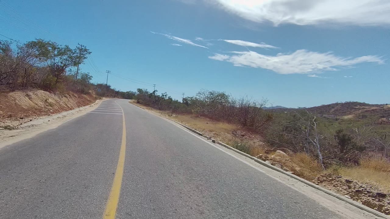 Forward road-level view of a two-lane road in East Cape Los Cabos, Baja California Sur, with dry roadside brush, low hills, scattered clouds, and no visible people.