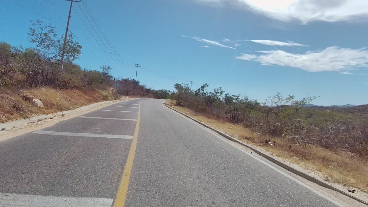 A two-lane road in East Cape Los Cabos, Baja California Sur, curves right past dry shrubs, rocky shoulders, utility lines, distant hills, and no visible people.