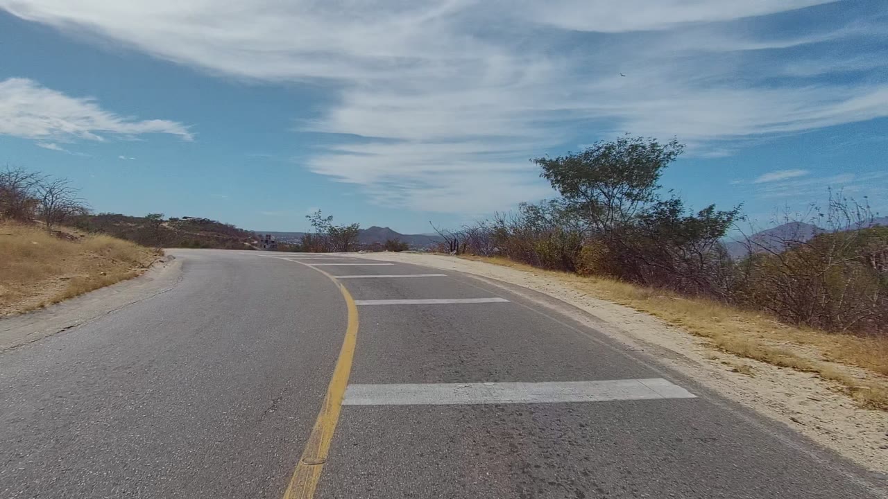 Opening view of a paved road curving left through dry East Cape Los Cabos hills, with scrub, small trees, blue sky, streaked clouds, and no visible people.