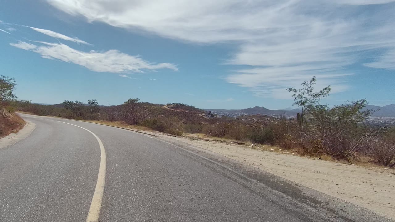 A later moment shows the East Cape Los Cabos road bending uphill between dry brush and sparse trees, with hazy hills on the horizon and no visible people.