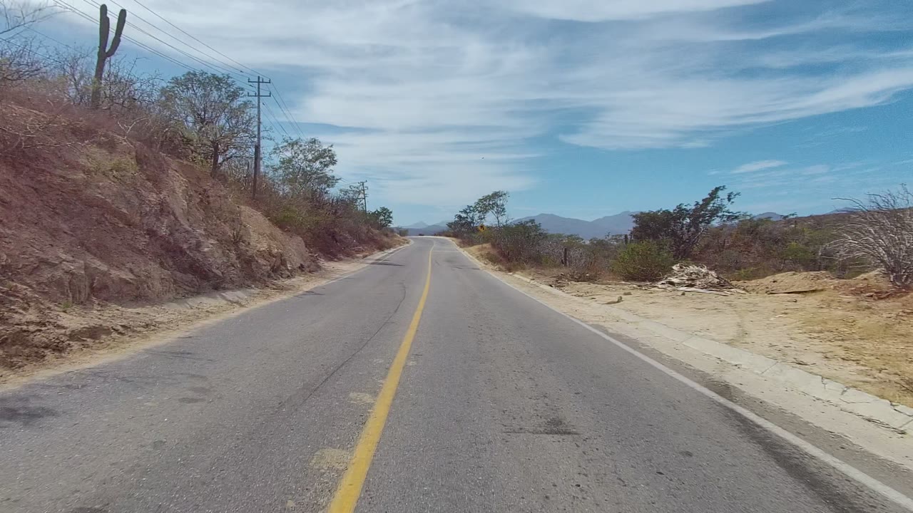 Forward view on an empty rural road in East Cape Los Cabos, with a yellow center line, dry roadside brush, cactus, utility poles, distant hills, scattered clouds, and no visible people.