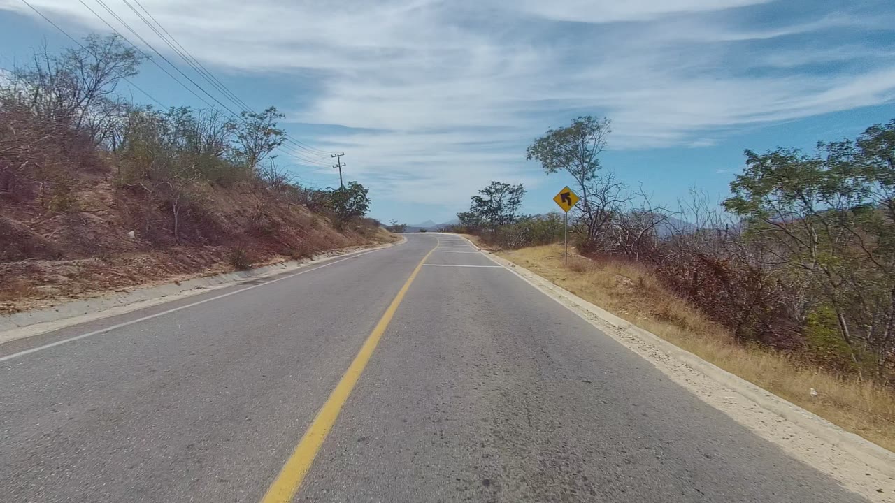 A forward road view in East Cape Los Cabos shows the yellow center line, sparse roadside trees, dry brush, power lines, a curve warning sign, distant hills, and no visible people.