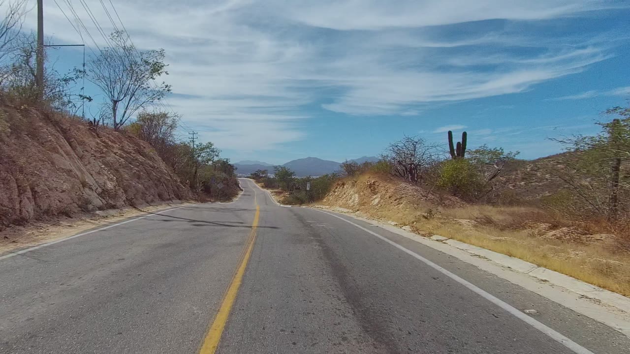 Forward view of a two-lane road in East Cape Los Cabos, Mexico, with a yellow center line, dry roadside slopes, sparse brush, cactus, distant mountains, and no visible people.