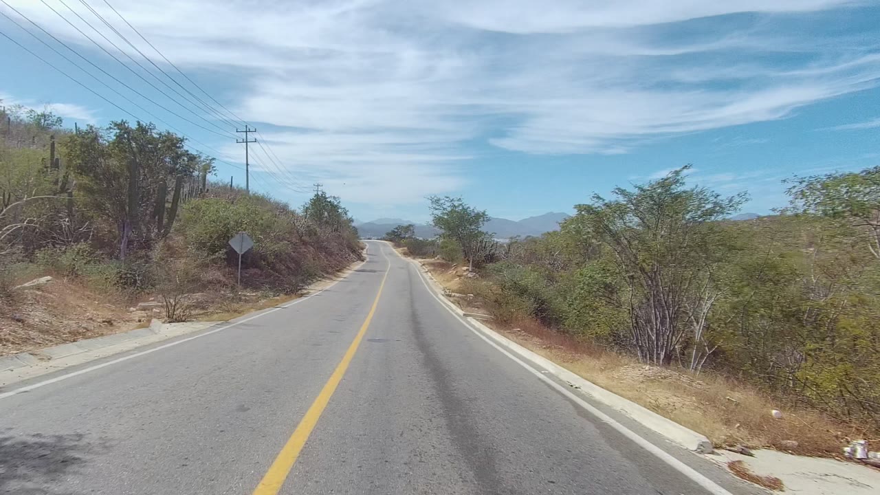 The road in East Cape Los Cabos, Mexico, continues toward low mountains under streaked clouds, with dry shoulders, roadside scrub, utility poles, a small road sign, and no visible people.