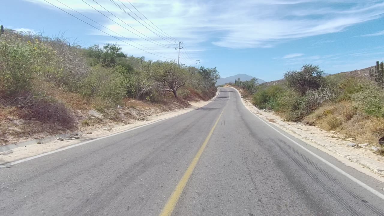 Forward view of a paved road in East Cape Los Cabos, Mexico, bordered by dry brush, small trees, utility lines, and low hills under a blue sky with no visible people.