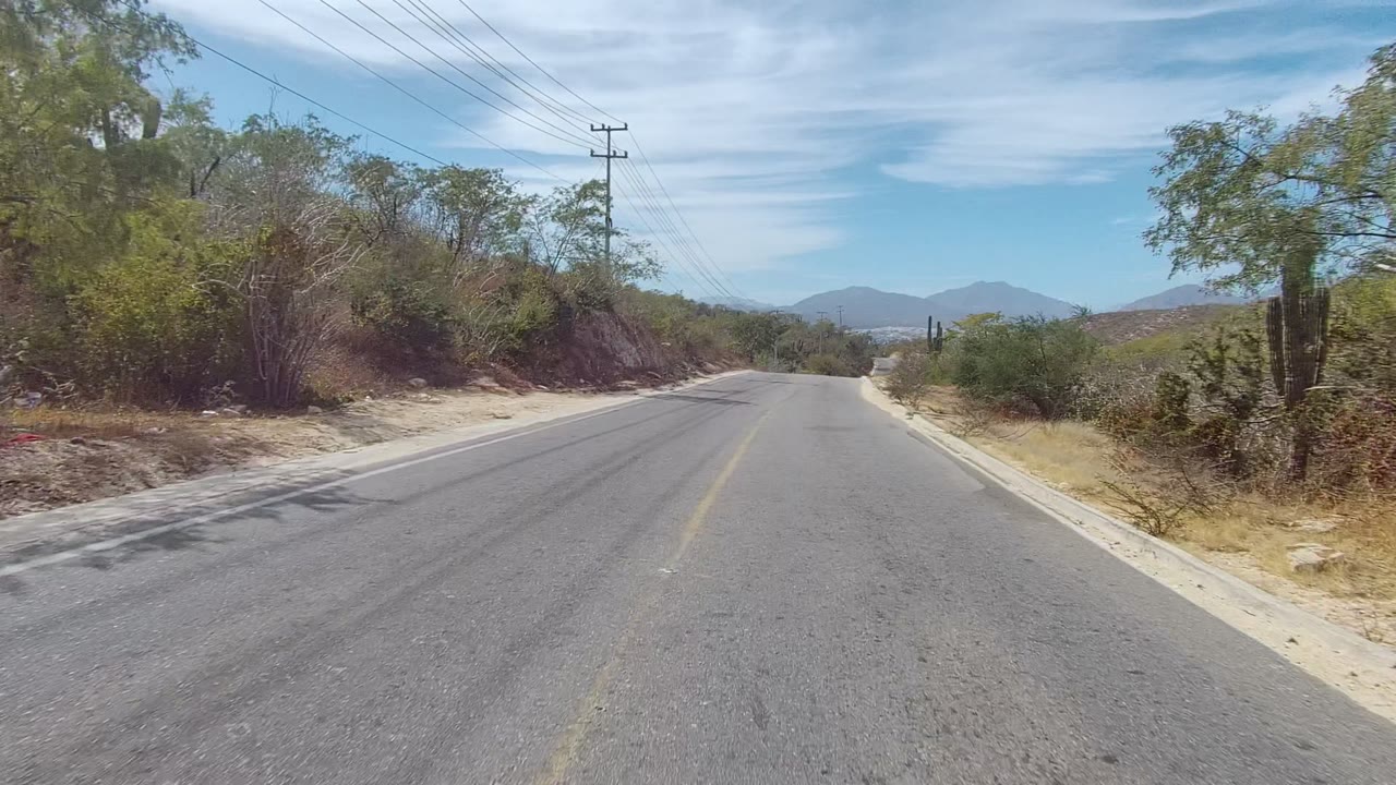 Forward view of a paved road in East Cape Los Cabos, Baja California Sur, with scrub along both shoulders, overhead power lines, distant mountains, thin clouds, and no visible people.