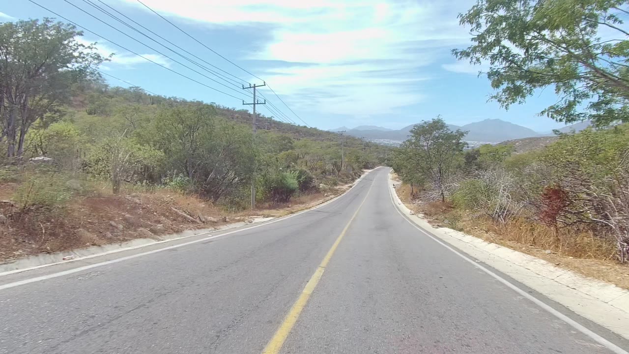 A two-lane asphalt road in East Cape Los Cabos runs between dry brush, trees, utility poles, distant hills, and a partly cloudy blue sky, with no visible people.