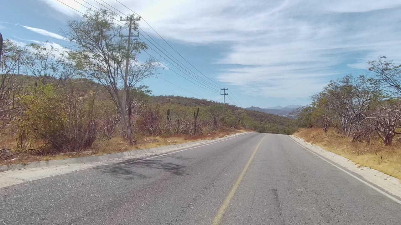 East Cape Los Cabos road view with a yellow center line, dry roadside brush, scattered cacti, utility wires, low hills, distant mountains, and no visible people.