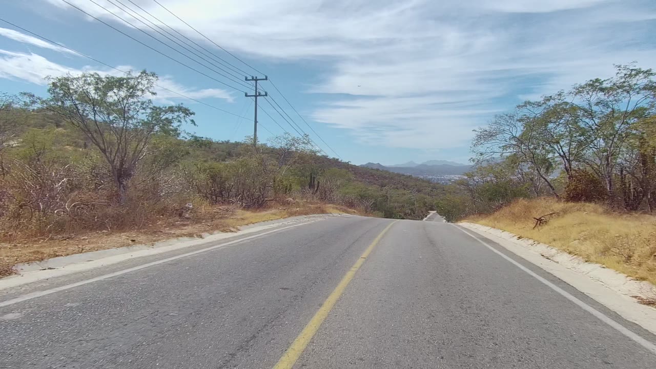 The East Cape Los Cabos road stretches ahead toward distant mountains, bordered by dry embankments, sparse green trees, scrub, utility lines, and no visible people.