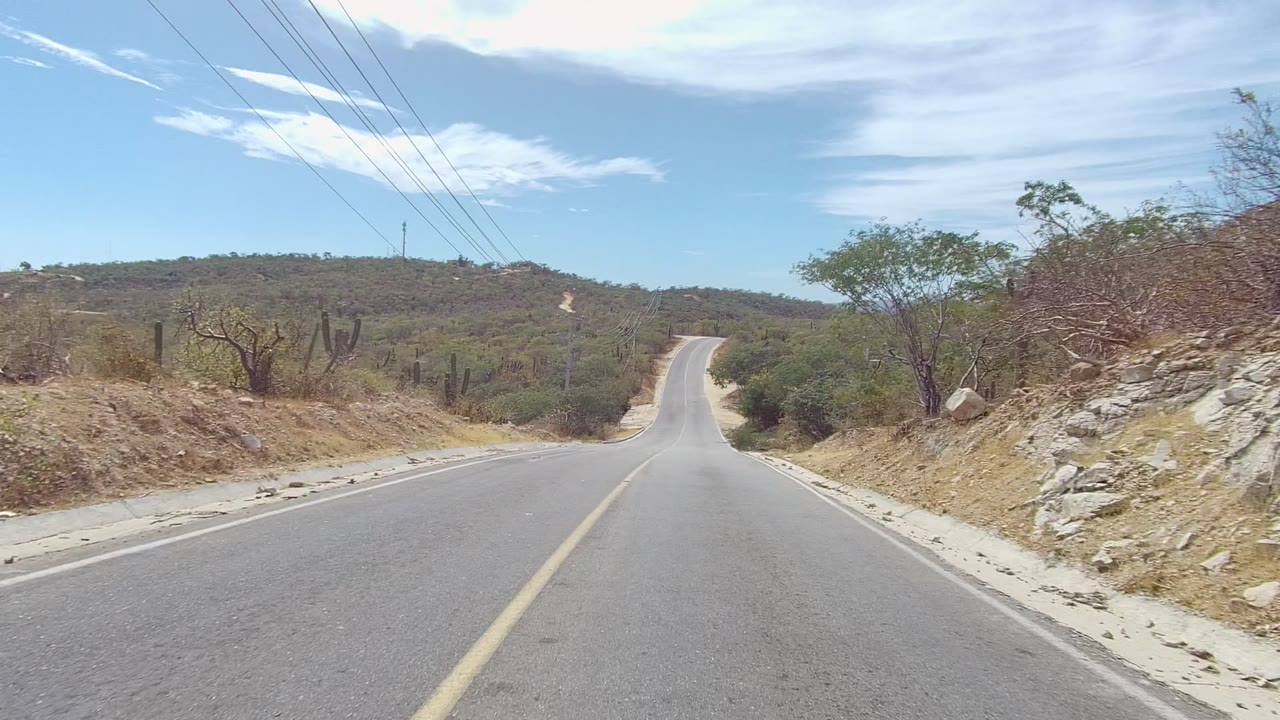 Paved road in East Cape Los Cabos, Baja California Sur, rising between dry roadside brush, cacti, power lines, and low hills with no visible people.