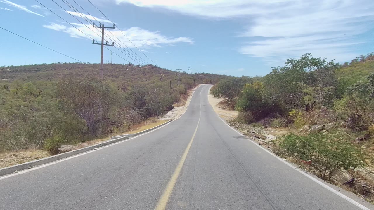 Paved road in East Cape Los Cabos, Baja California Sur, leading toward a small rise with a utility pole, power lines, dry shrubs, trees, and no visible people.
