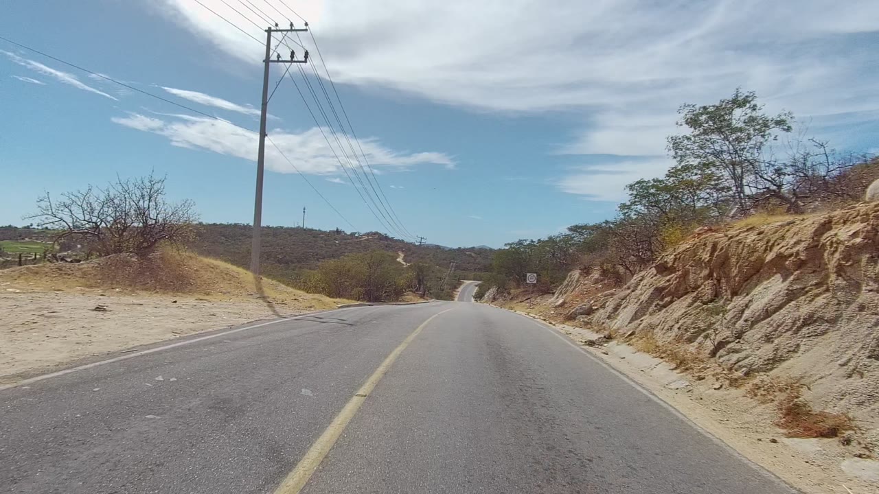 A two-lane road in East Cape Los Cabos runs through dry terrain with a utility pole on the left, rocky bank on the right, blue sky overhead, and no visible people.