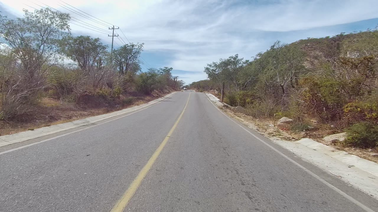 Forward road-level view of a two-lane road in East Cape Los Cabos, Mexico, bordered by dry shrubs, trees, cacti, utility lines, and no visible people.