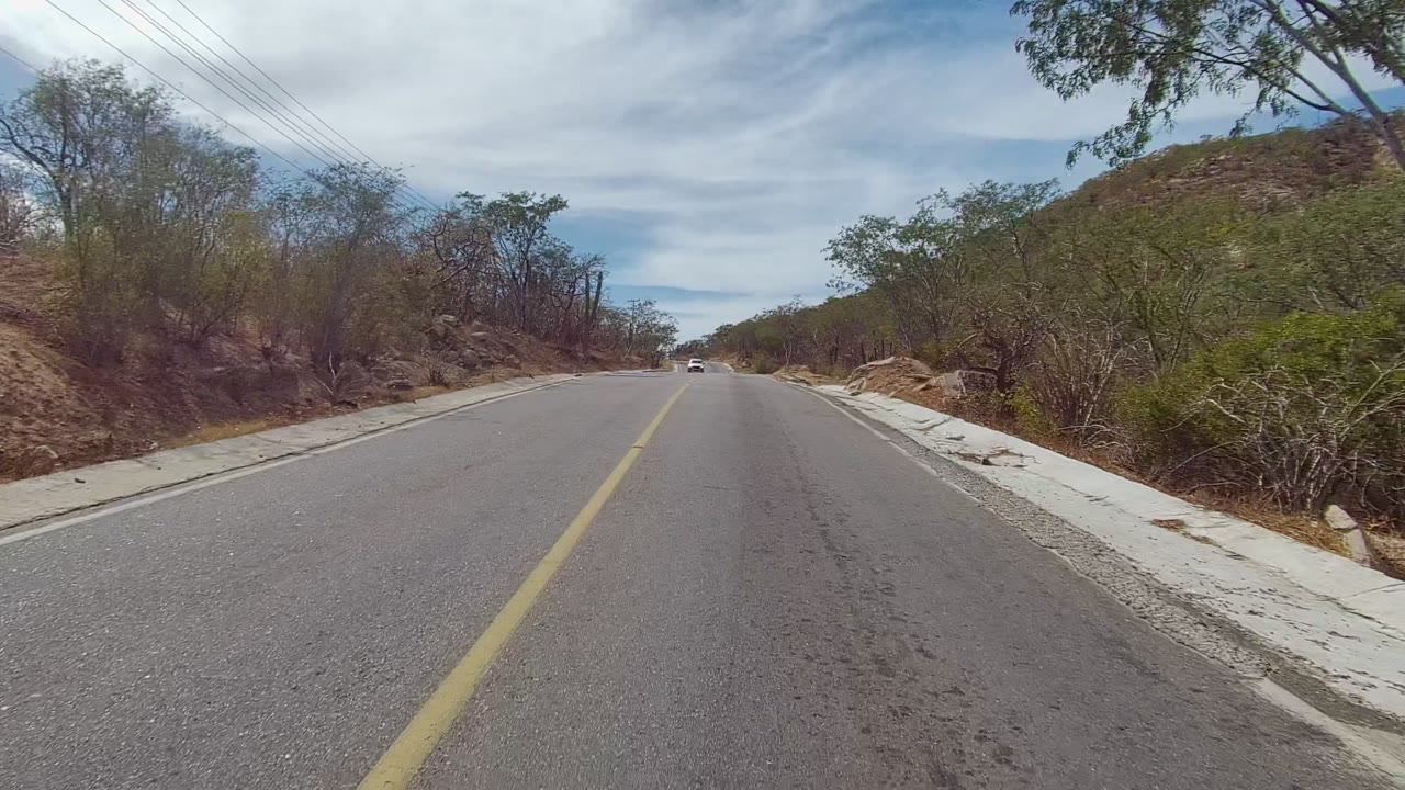 A distant oncoming vehicle appears on the two-lane East Cape Los Cabos road, with dry brush, cacti, low hills, utility lines, and no visible people.