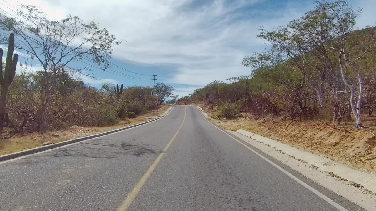 A two-lane road in East Cape Los Cabos, Baja California Sur, runs uphill between dry brush, cactus, utility lines, and low hills, with no visible people.