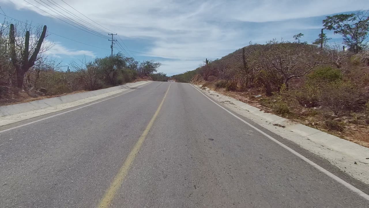 East Cape Los Cabos paved road seen from road level with a yellow center line, sandy shoulder, cacti, dry brush, low hillside, utility lines, partly cloudy sky, and no visible people.