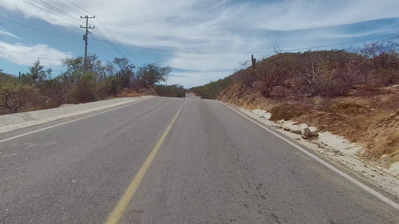 Empty paved road curving past dry brush and a low hillside
