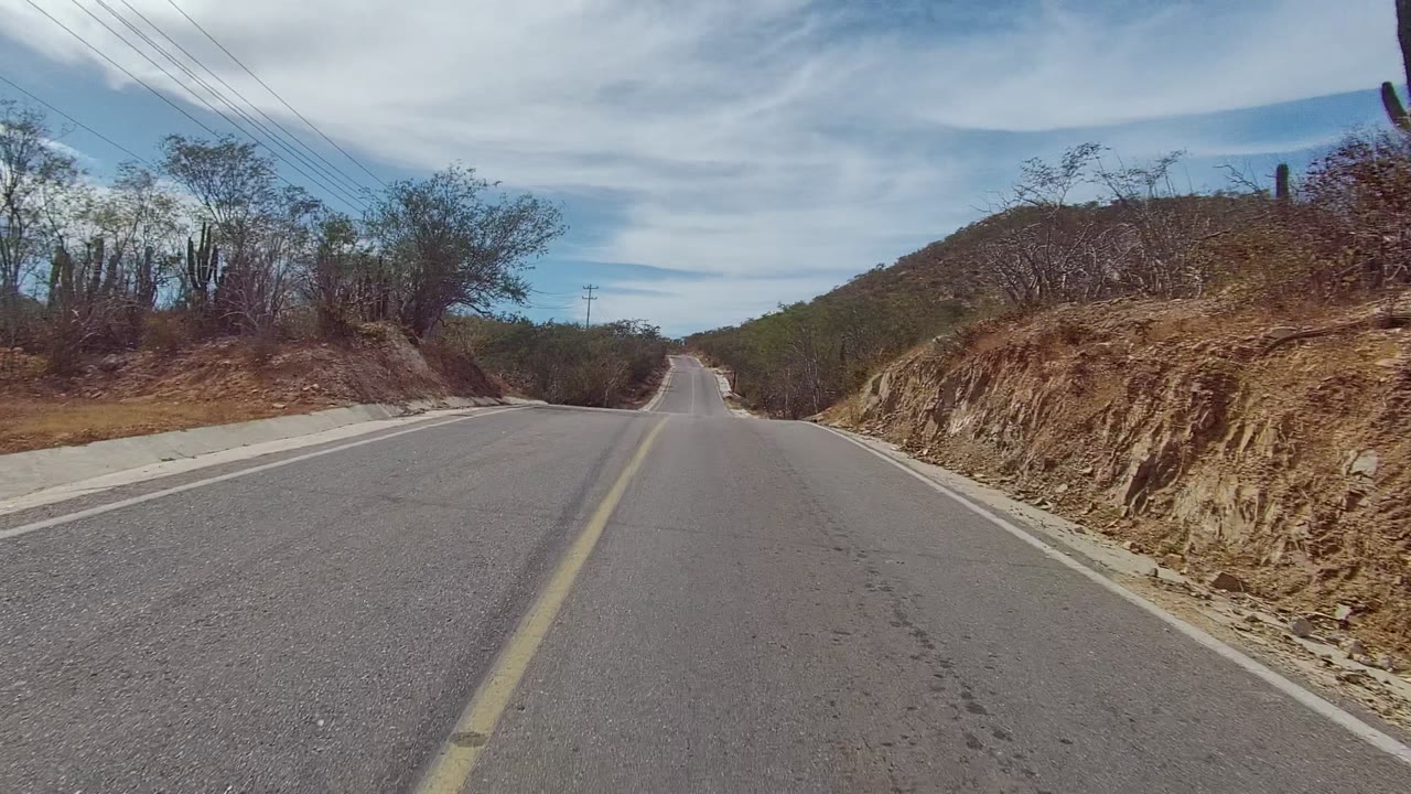 East Cape Los Cabos road framed by a cut dirt bank on the right, dry brush and cacti on the left, a yellow center line, pale road shoulders, utility lines overhead, layered clouds, and no visible people.