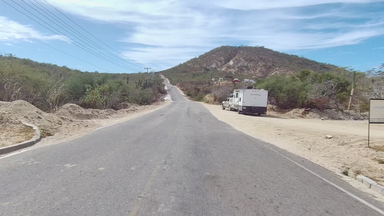 A paved road in East Cape Los Cabos runs toward a low hill, with sandy shoulders, dry brush, utility lines, parked white vehicles on the right, and no visible people.