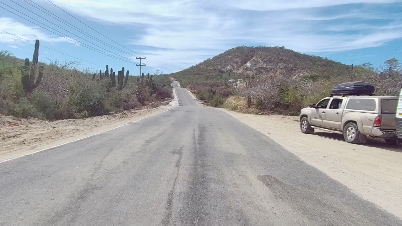 Desert road with a signboard, cacti, utility poles, and a hill in the distance.