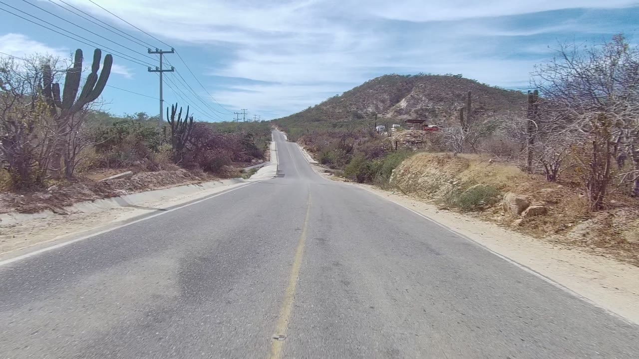 A light-colored pickup truck sits on the right shoulder of an East Cape Los Cabos desert road, with tall cacti, scrub, utility lines, distant hills, and no visible people.