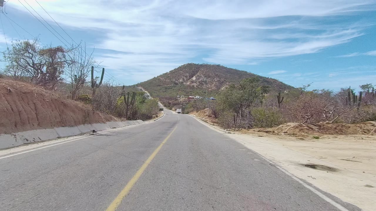 Paved road in East Cape Los Cabos, Baja California Sur, Mexico, with a yellow center line, dry roadside brush, cacti, a low hill ahead, thin clouds, and no visible people.