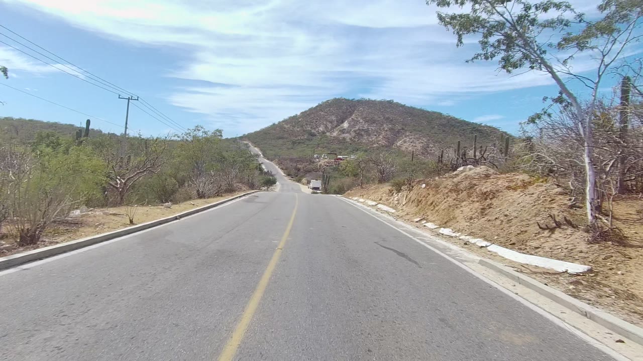 Paved road in East Cape Los Cabos with dry brush on both sides, utility poles and overhead lines at right, a rounded hill in the distance, and no visible people.