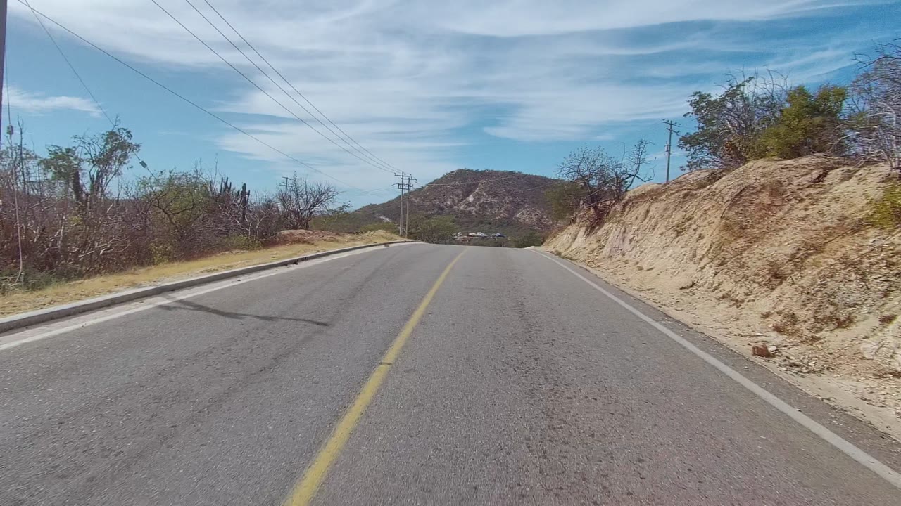 Opening view of a paved road in East Cape Los Cabos, Baja California Sur, with a yellow center line, sandy cut bank, sparse roadside trees, distant hill, and no visible people.