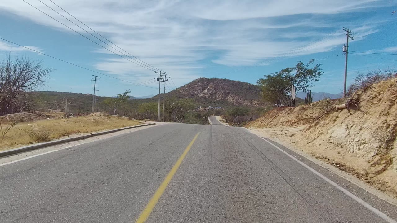 Two-lane road curving through scrub with poles and hills beyond.