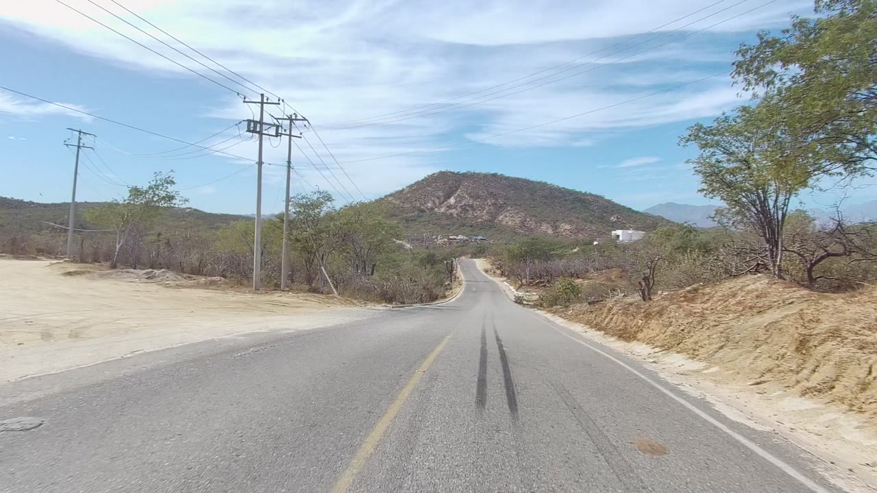 East Cape Los Cabos road frame with the pavement leading downhill, dry brush on both sides, a rounded rocky hill ahead, utility poles at the roadside, and no visible people.