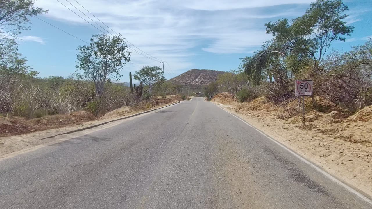 Opening view of a two-lane rural road in East Cape Los Cabos, Mexico, with a roadside 50 km/h sign, sandy shoulders, scrub vegetation, cacti, power lines, low hills, and no visible people.