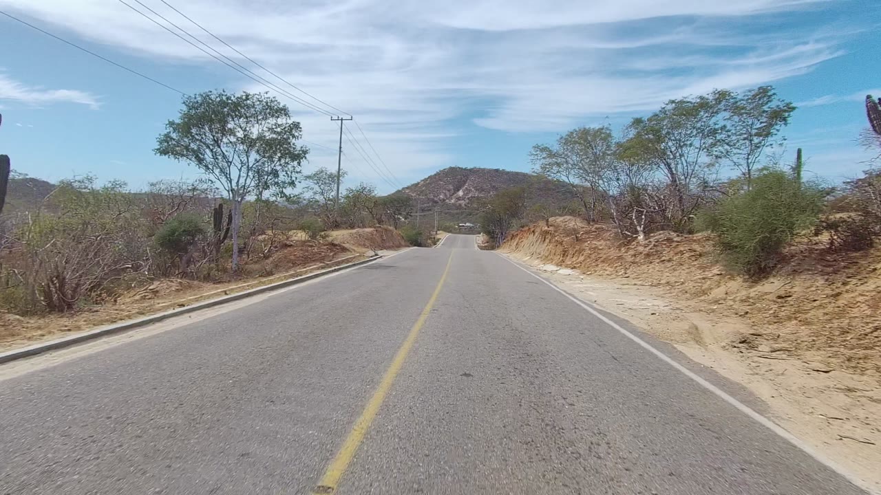 Road curving slightly through dry scrub with a yellow center line and low hills ahead.